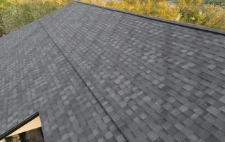 Aerial view of a house roof covered with dark gray asphalt shingles, surrounded by trees with green and yellow foliage.