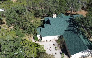 Aerial view of a large house with a green roof surrounded by trees, with a gravel area and some outdoor furniture visible in the yard.