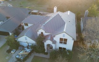 Aerial view of a two-story white house with a metal roof, flanked by neighboring houses, with a construction waste bin and debris in the driveway.