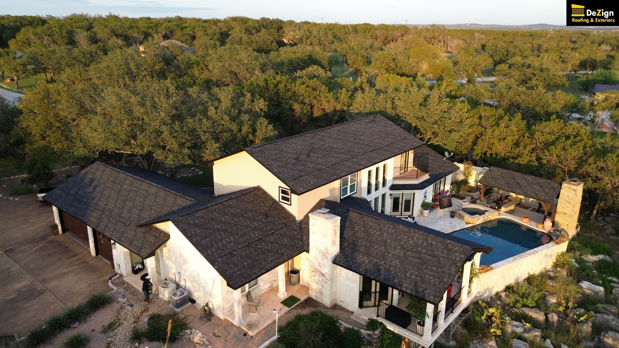 Aerial view of a large house with a dark shingle roof, a swimming pool, patio area, garage, and surrounding trees. DeZign Roofing & Exteriors logo appears in the top right corner.