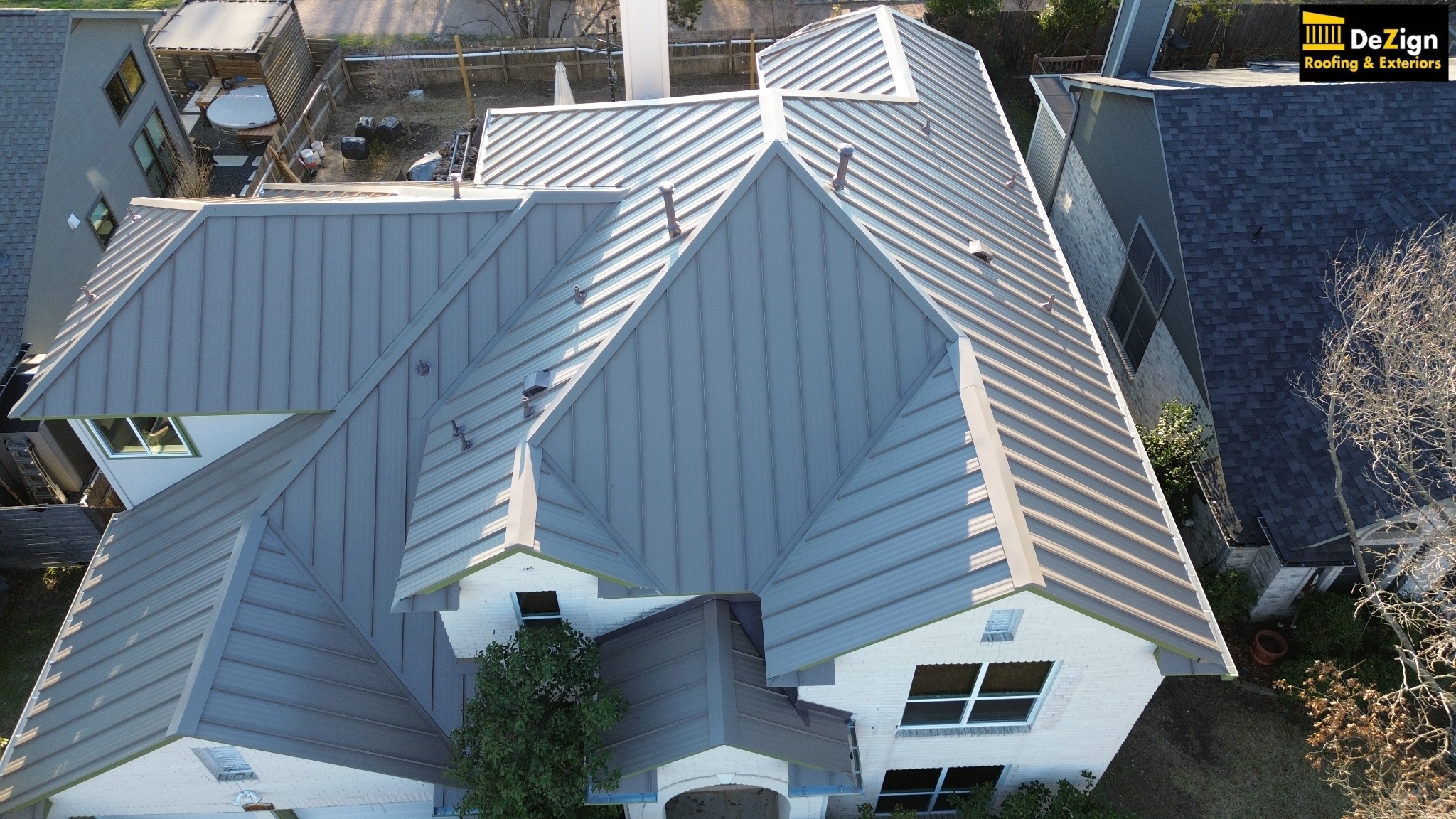 Aerial view of a modern house with a gray metal roof featuring multiple ridges and slopes, surrounded by neighboring buildings and trees.